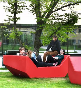 Junge Menschen auf großen roten Kunststoff-Möbeln unter Bäumen am Campus Friedrich Streib der Hochschule Coburg..