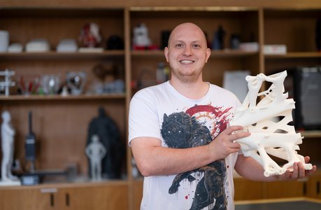 Felix Felgenträger steht in einem Labor der Hochschule Coburg vor einem hölzernen Regal mit 3D-gedruckten Exponaten.