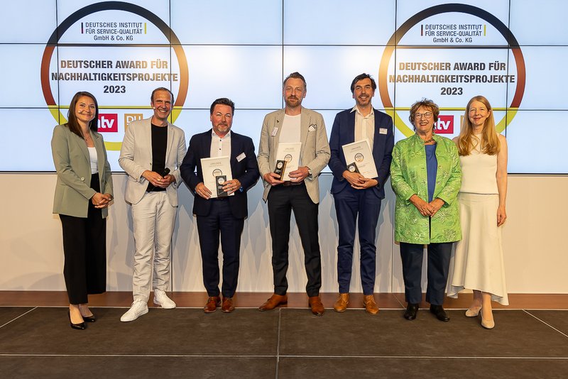 Eine Gruppe von sieben Personen der Hochschule Coburg steht auf einer Bühne, einige halten stolz Auszeichnungen in den Händen. Sie posieren vor einer Leinwand mit dem Logo des „Deutschen Awards für Nachhaltigkeitsprojekte 2023“, sind in Business-Kleidung gekleidet und lächeln für das Foto.