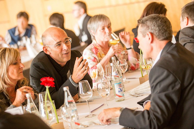 A group of people from Hochschule Coburg sits at a table engaged in conversation. The table is set with wine glasses, water bottles, and red gerbera daisy centerpieces. The atmosphere appears cordial and social in a well-lit room.