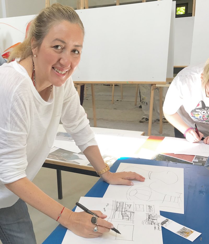 A person is smiling while drawing on a large sheet of paper at a Hochschule Coburg classroom table. Behind them, easels with blank canvases await inspiration. Another person is partially visible, deeply focused on their drawing in this creative studio environment.