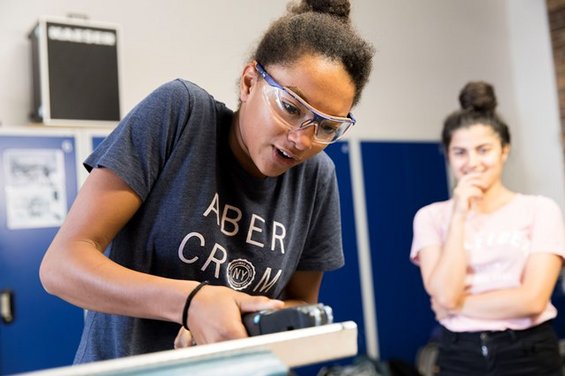 Eine Person mit Schutzbrille und Abercrombie-Shirt bearbeitet mit einem Werkzeug ein Stück Holz und verfeinert dabei wahrscheinlich die an der Hochschule Coburg erlernten Fähigkeiten. Eine andere Person steht in der Nähe, schaut zu und lächelt, in einem Raum mit blauen Schließfächern.