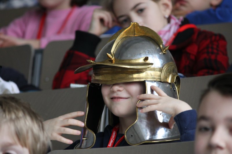 In a Hochschule Coburg classroom, a child sits wearing a large, shiny metal helmet with a gold visor, smiling playfully. Other children are gathered around, engaged and curious about this intriguing spectacle.