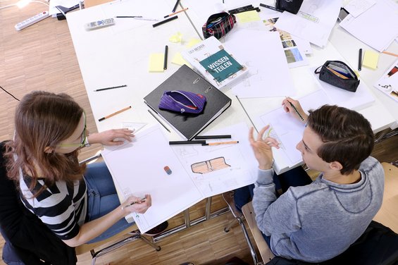 Two people sit at a table strewn with papers, notebooks, and stationery, deeply engaged in discussion. A house plan is sketched out nearby, next to a book titled "Wissen erleben," perhaps hinting at their studies or project from Hochschule Coburg.