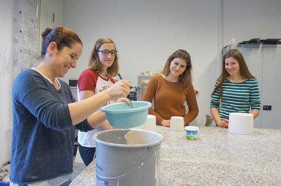 Eine Frau mit dunklem Haar bereitet eine Mischung in einer blauen Schüssel zu, während drei junge Frauen von der Hochschule Coburg zuschauen. In dem Raum mit einem grauen Tisch und Regalen sind sie vertieft in eine Tätigkeit, möglicherweise Töpfern oder Basteln.