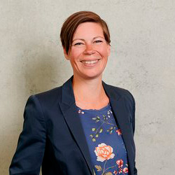 A smiling person with short brown hair, wearing a navy blazer over a floral top, stands confidently against a light gray background, embodying the professional spirit of Hochschule Coburg.