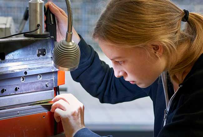 A young person with blonde hair, tied in a ponytail, carefully operates a piece of machinery under the focused light at Hochschule Coburg. They appear concentrated on the task, adjusting a component with precision. The background is blurred, highlighting the machinery.