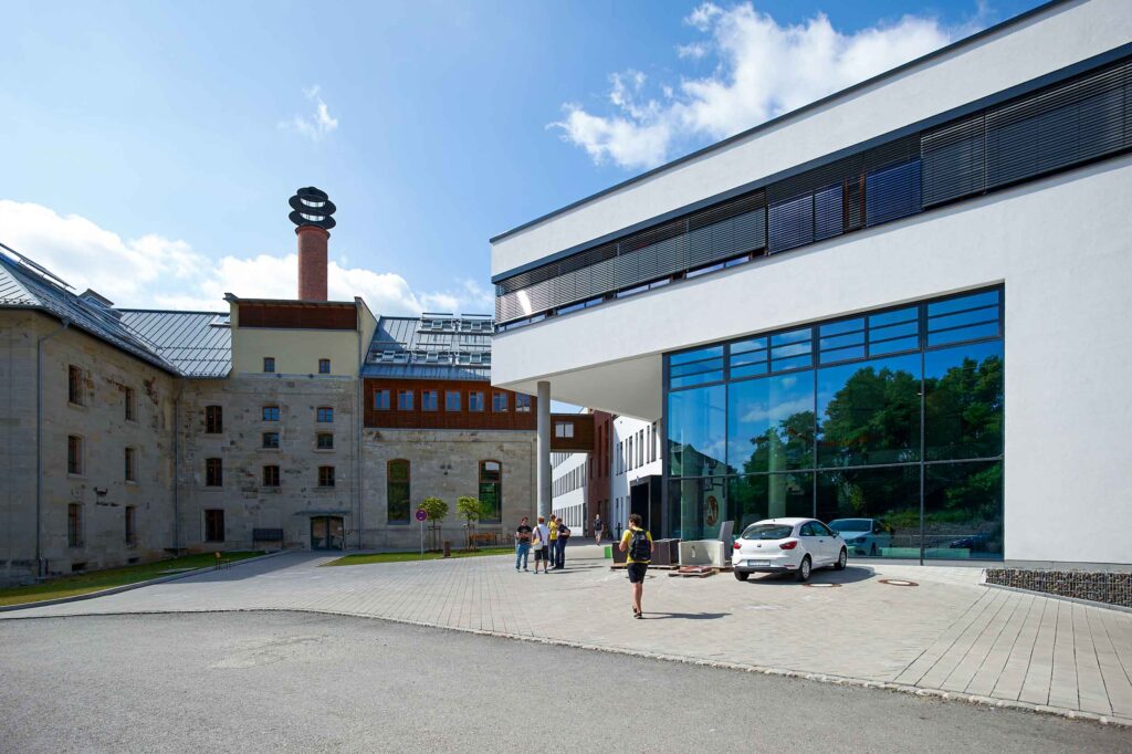 The contemporary building with large windows, next to the historic structure featuring a brick chimney, reflects the architectural harmony found at Hochschule Coburg. On this sunny day, a few people stroll by while a parked car sits in the foreground.