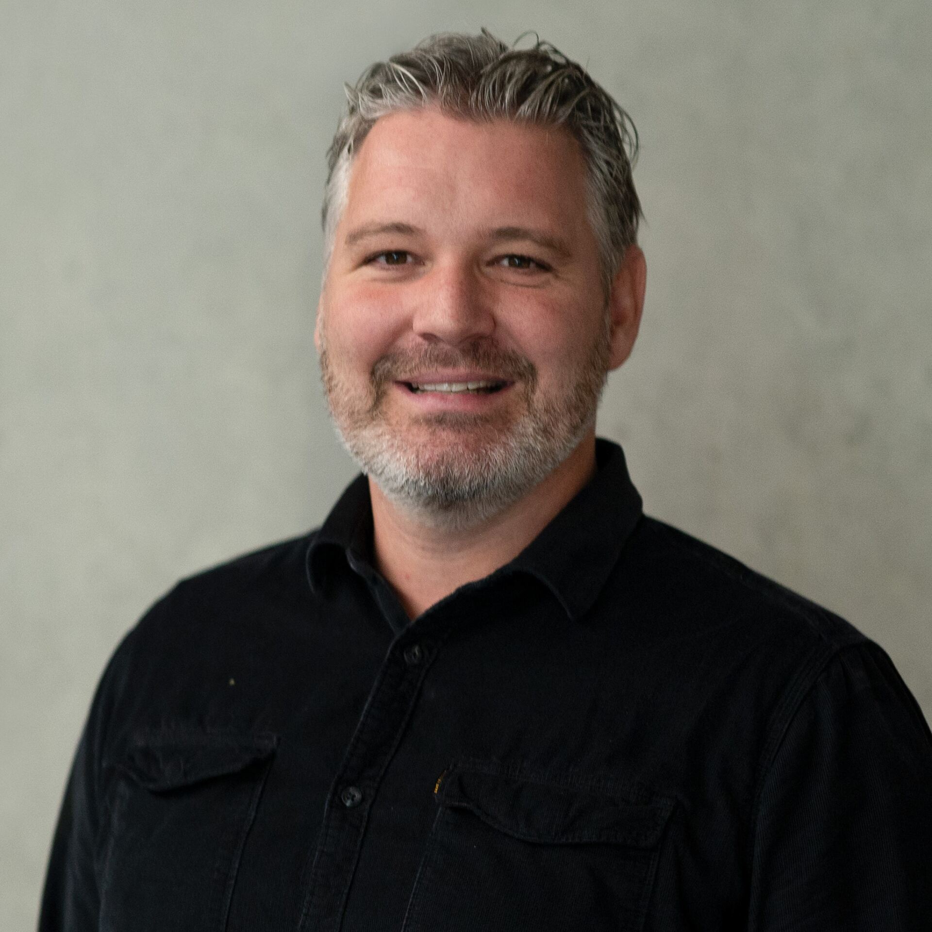 Timo Bertocchi, with his short gray hair and beard, is smiling warmly in a black shirt. The neutral gray background complements his friendly demeanor perfectly.