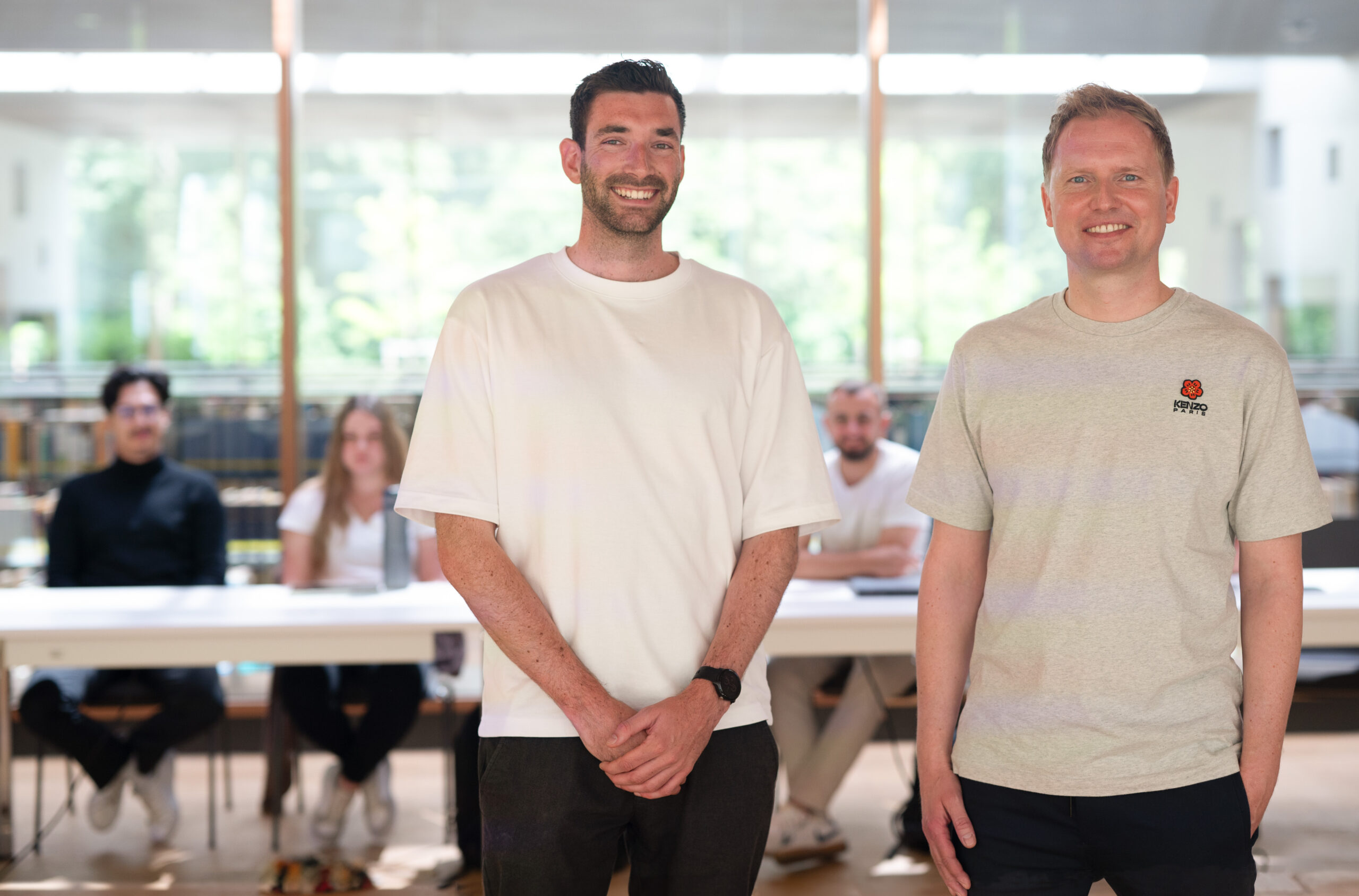 Two men, smiling and standing in front of a table, are enjoying a relaxed and informal moment at Hochschule Coburg. Behind them, four seated people are blurred, while a large window reveals the greenery outside.