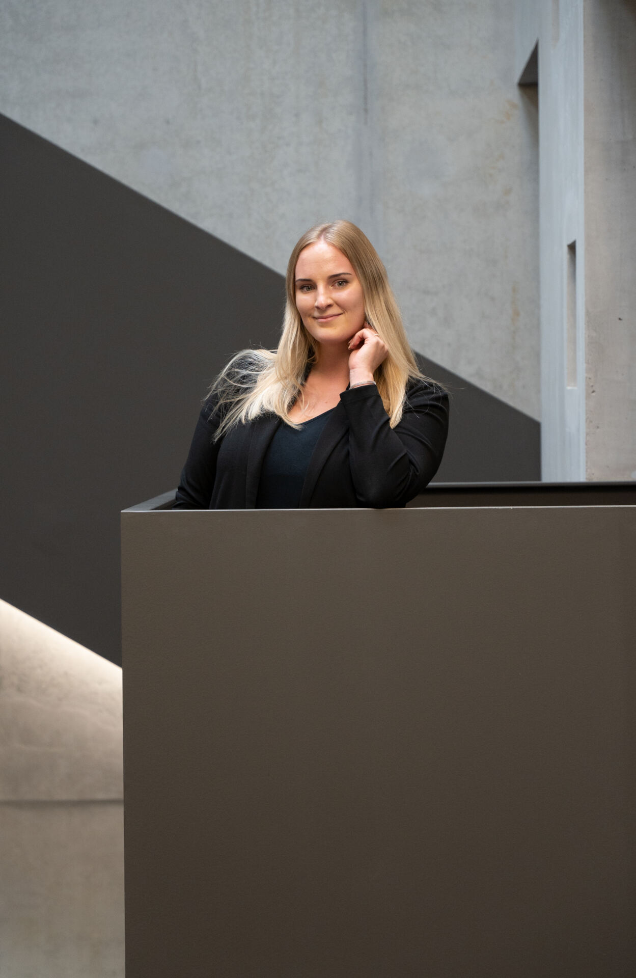 Eine Frau mit langen blonden Haaren steht und lehnt an einem dunklen Geländer der Hochschule Coburg. Sie trägt einen schwarzen Blazer und lächelt sanft. Im Hintergrund ist moderne, geometrische Architektur mit Beton und diagonalen Linien zu sehen.