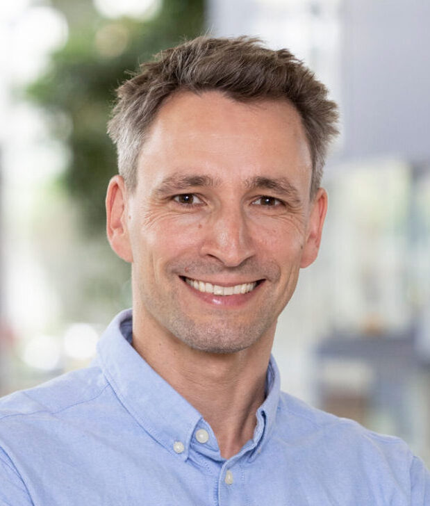 Smiling warmly at the camera, a person with short grayish hair and a light blue button-up shirt stands in front of a softly blurred indoor setting, suggesting they might be on campus at Hochschule Coburg, where hints of greenery and light create an inviting atmosphere.