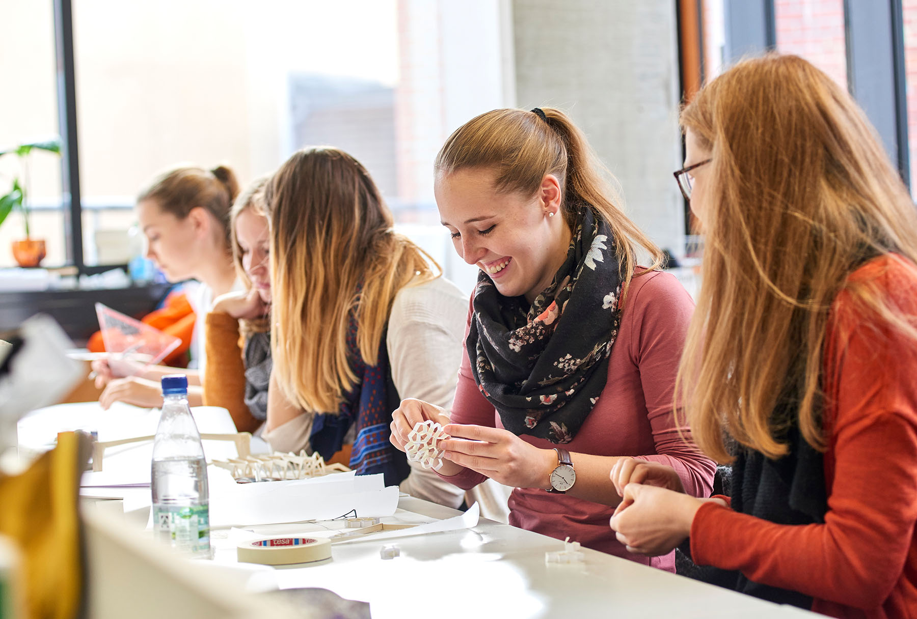 In einem hellen Raum der Hochschule Coburg beschäftigt sich eine Gruppe junger Frauen mit Bastelarbeiten. Eine Frau strahlt, während sie ein komplexes geometrisches 3D-Modell in den Händen hält. Der Tisch ist voll mit Materialien und Werkzeugen, und das Sonnenlicht fällt durch die hohen Fenster und schafft eine inspirierende Atmosphäre.