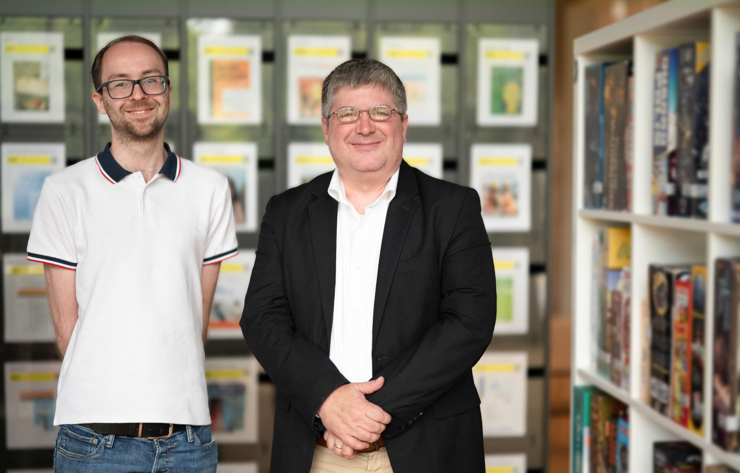 Two men stand side by side, smiling in a well-lit room at Hochschule Coburg. One wears a white polo shirt and jeans, while the other sports a black blazer with a white shirt. Behind them are shelves filled with board games and a display of colorful magazine covers.