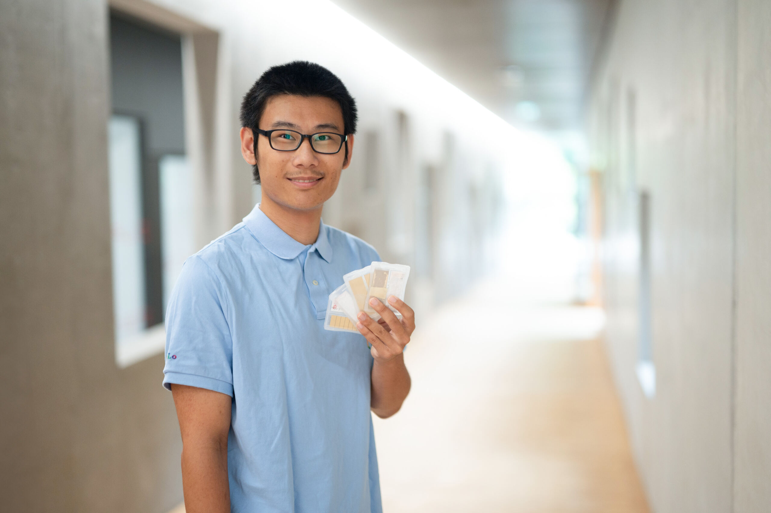 Eine vor Begeisterung strahlende Person im blauen Hemd hält einen Fächer aus Papier in der Hand und steht im hellen Flur der Hochschule Coburg.