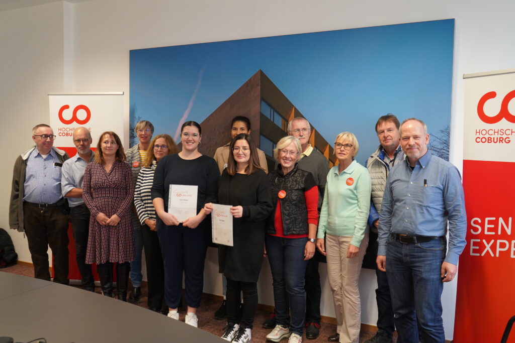 Eine Gruppe von Menschen steht drinnen vor einem großen Foto eines Gebäudes, auf beiden Seiten sind zwei rote Banner der Hochschule Coburg zu sehen. Zwei Personen halten stolz Zertifikate in den Händen und alle lächeln herzlich.