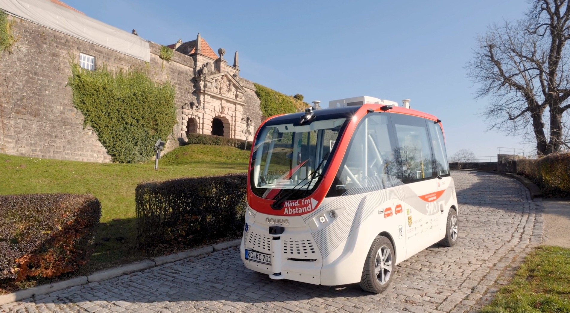 Ein kleines, fahrerloses Shuttle mit rot-weißem Design steht auf einem Kopfsteinpflasterweg in der Nähe eines alten Steingebäudes der Hochschule Coburg. Der Himmel ist klar und Bäume mit spärlichem Laub säumen die Straße und fangen die Harmonie von Tradition und Innovation ein.