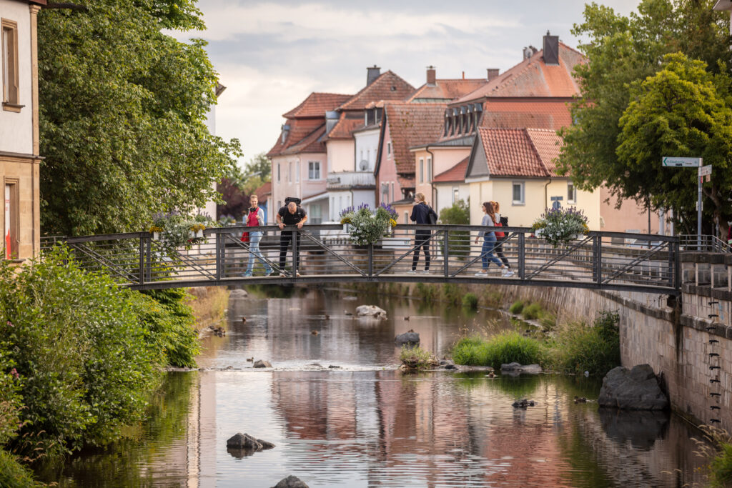 Eine kleine Gruppe von Menschen steht auf einer dekorativen Brücke über einem ruhigen Fluss in einem malerischen Dorf in der Nähe der Hochschule Coburg. Die Straße ist gesäumt von charmanten, farbenfrohen Häusern und üppige grüne Bäume rahmen die Szene unter einem bewölkten Himmel ein.