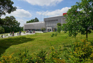 A modern building with a sleek exterior, reminiscent of Hochschule Coburg's architectural elegance, stands surrounded by greenery and trees. People are sitting and strolling on the grassy lawn in front. The sky is partly cloudy, creating a bright and tranquil atmosphere.