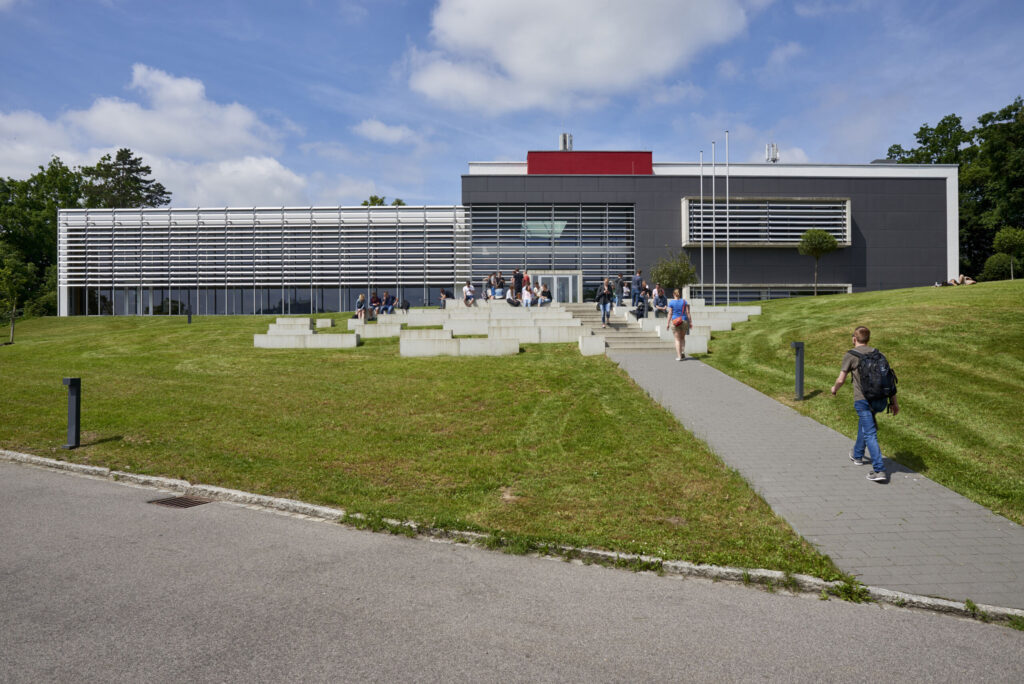 Der Campus der Hochschule Coburg besteht aus einem modernen Universitätsgebäude mit großen Fenstern und geometrischem Design, umgeben von einer üppigen Rasenfläche. Unter einem teilweise bewölkten Himmel schlendern Studenten den Weg zum Eingang entlang.