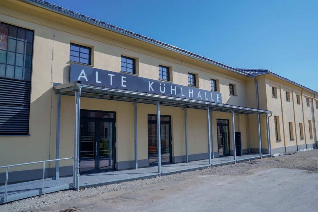 Ein gelbes Gebäude mit einem Metallvordach und der Aufschrift „ALTE KÜHLHALLE“, das an den Architekturstil der Hochschule Coburg erinnert, verfügt über große Fenster und Türen. Das Gebäude hat ein Flachdach unter einem klaren blauen Himmel, mit Stufen und einer Rampe, die zum Eingang führen.