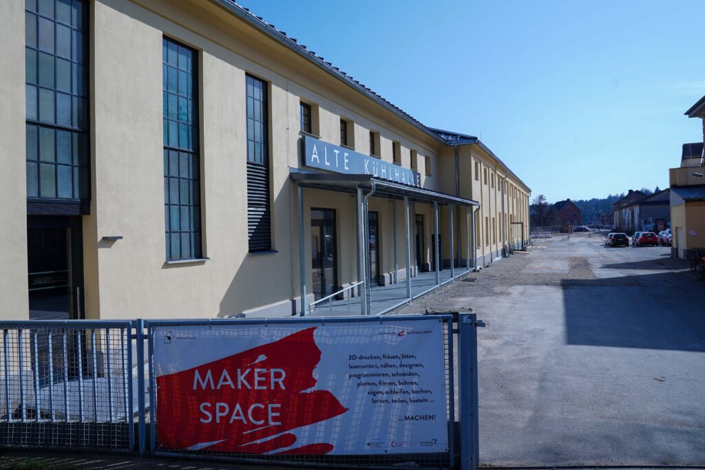 Außenansicht eines langen, beigen Gebäudes mit großen Fenstern und einem Schild mit der Aufschrift „Alte Kühlhalle“, Teil des lebendigen Campus der Hochschule Coburg. Ein Banner an einem Zaun im Vordergrund wirbt für einen „Maker Space“, in dem kreative Aktivitäten aufgelistet sind. Ein klarer, sonniger Tag mit blauem Himmel verstärkt die einladende Atmosphäre.