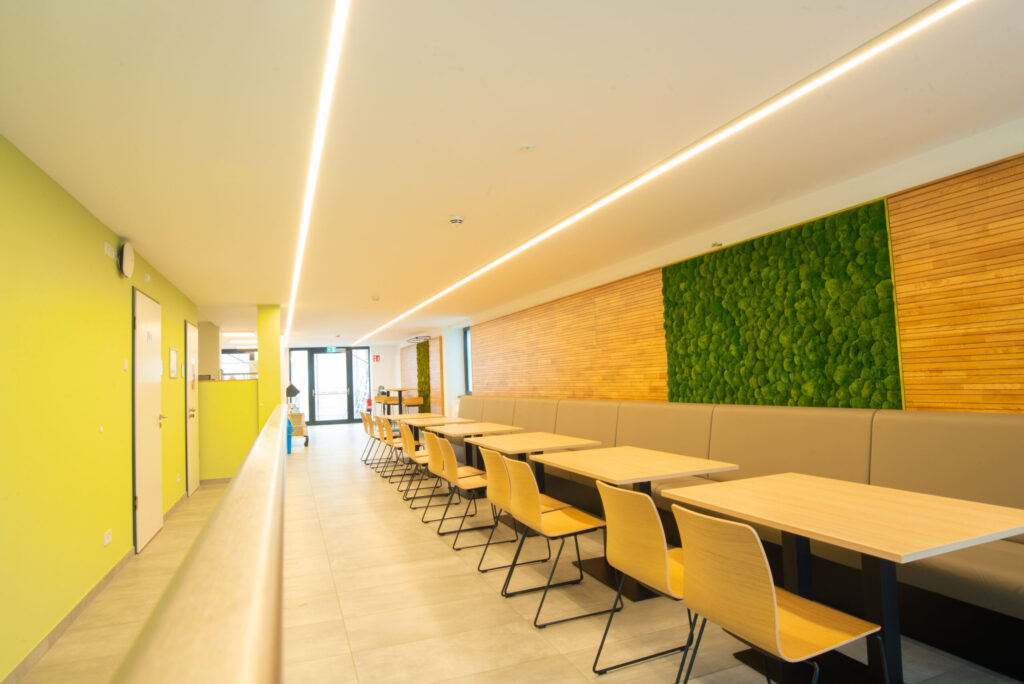 The modern cafeteria at Hochschule Coburg features long tables, light wooden chairs, and green walls with a vertical plant display. Bright natural light pours in through large windows, giving the space a sleek, contemporary look that inspires both students and faculty alike.