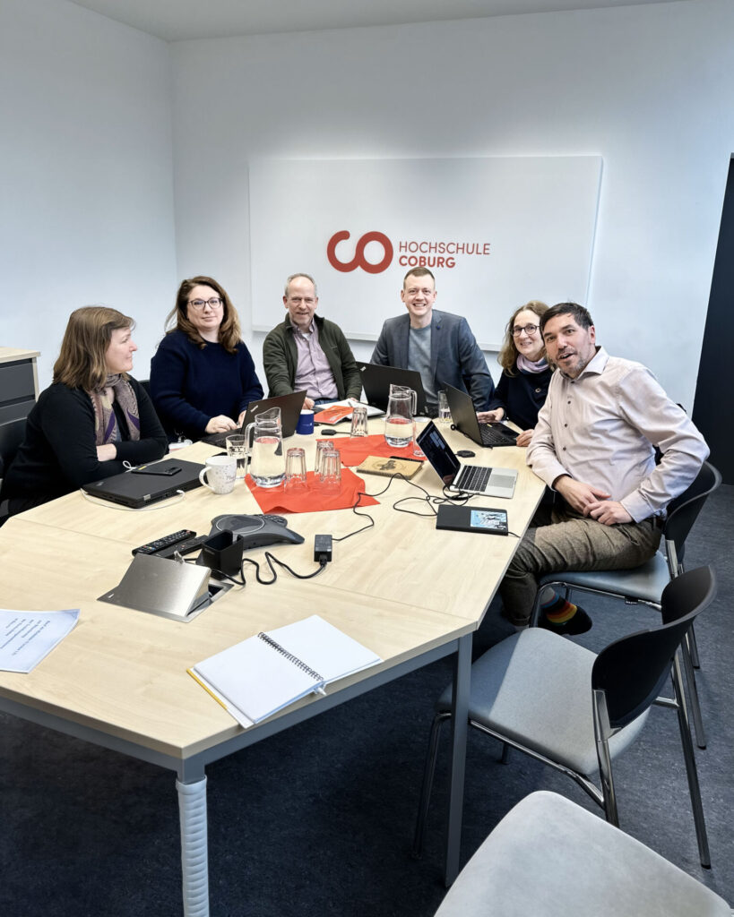 A group of six people sits around a conference table with laptops and documents at Hochschule Coburg. The room reflects a friendly and collaborative atmosphere.