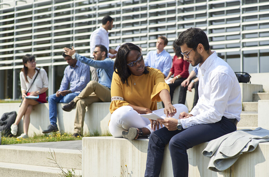 Eine Gruppe unterschiedlicher Menschen sitzt auf Außentreppen in einem modernen, urbanen Ambiente der Hochschule Coburg. Im Vordergrund bespricht eine Frau in einem gelben Oberteil und einer Sonnenbrille Papiere mit einem Mann in einem weißen Hemd, während andere sich unterhalten oder sich umsehen und die lebhafte Atmosphäre aufsaugen.