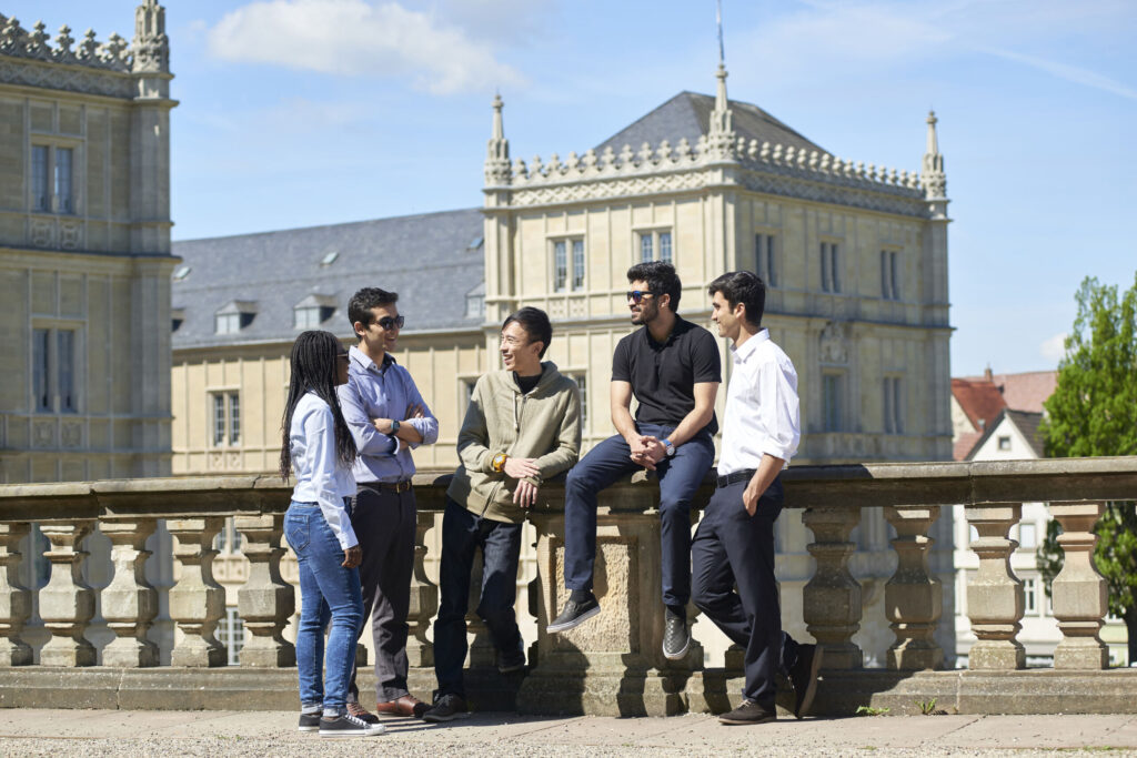 Fünf Personen unterhalten sich draußen auf einer Steinterrasse der Hochschule Coburg, mit historischen Gebäuden und einem klaren blauen Himmel im Hintergrund. Die Gruppe wirkt entspannt, in legerer und elegant-legerer Kleidung gekleidet.