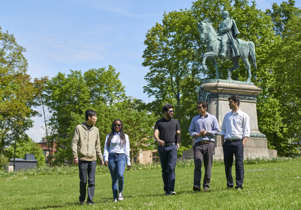 Eine Gruppe von fünf Personen, vermutlich Studierende der Hochschule Coburg, schlendern gemeinsam über eine Wiese in einem Park. Im Hintergrund ist eine Statue eines Reiters zu sehen. Unter klarem Himmel und umgeben von ruhigen Bäumen unterhalten sie sich angeregt.