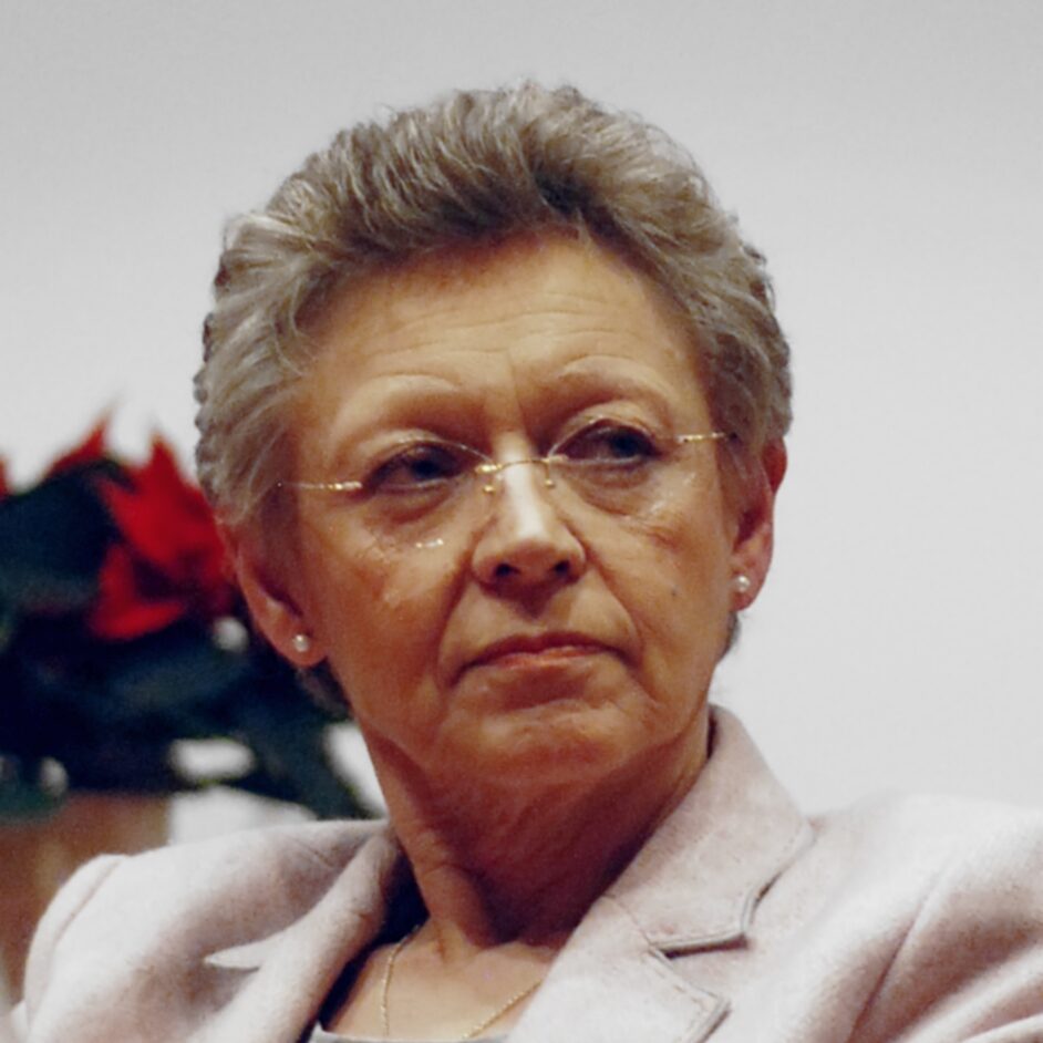 An older person with short gray hair and glasses wears a light-colored jacket, gazing thoughtfully to the side. Behind them, a plant with red flowers is softly blurred. The scene evokes the quiet, scholastic atmosphere reminiscent of Hochschule Coburg.