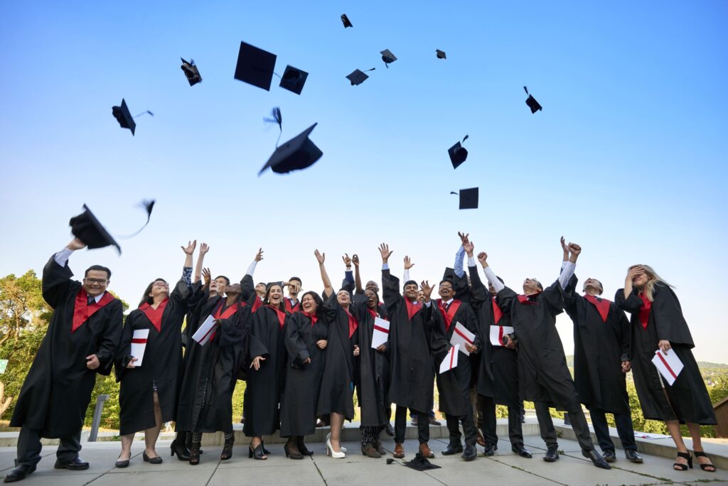 Eine Gruppe von Absolventen der Hochschule Coburg in schwarzen Talaren und Hüten feiert im Freien und wirft ihre Hüte voller Freude in den strahlend blauen Himmel. Vor dem klaren Hintergrund rahmen Bäume die Szene ein und strahlen Begeisterung aus.