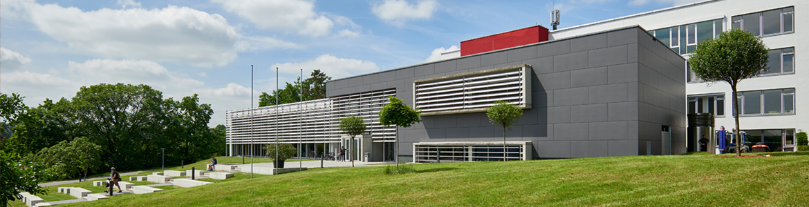 Ein modernes Gebäude mit einer eleganten grau-weißen Außenfassade, die an den zeitgenössischen Stil der Hochschule Coburg erinnert, verfügt über horizontale Lamellen und große Fenster. Die Umgebung besticht durch grünes Gras, kleine Bäume und ein paar Menschen, die unter einem blauen Himmel mit vereinzelten Wolken spazieren.