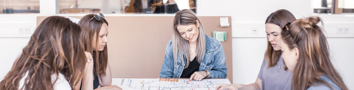 Fünf Frauen sitzen an einem Tisch in der Hochschule Coburg und diskutieren. Sie blicken auf ein großes Blatt mit bunten Linien und Markern. Die Atmosphäre scheint ein lockeres Meeting oder eine Brainstorming-Sitzung in einem gut beleuchteten Raum zu sein.