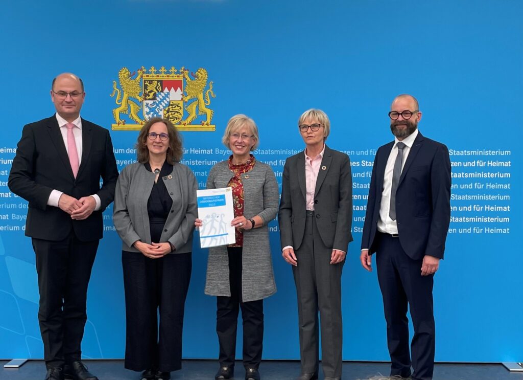 Eine Gruppe von fünf Personen in formeller Kleidung steht vor einem blauen Hintergrund mit dem bayerischen Wappen bei einer Veranstaltung der Hochschule Coburg. Die zentrale Figur hält ein Dokument in der Hand, und alle posieren anmutig und fangen die Formalität und das Prestige dieses offiziellen Anlasses ein.