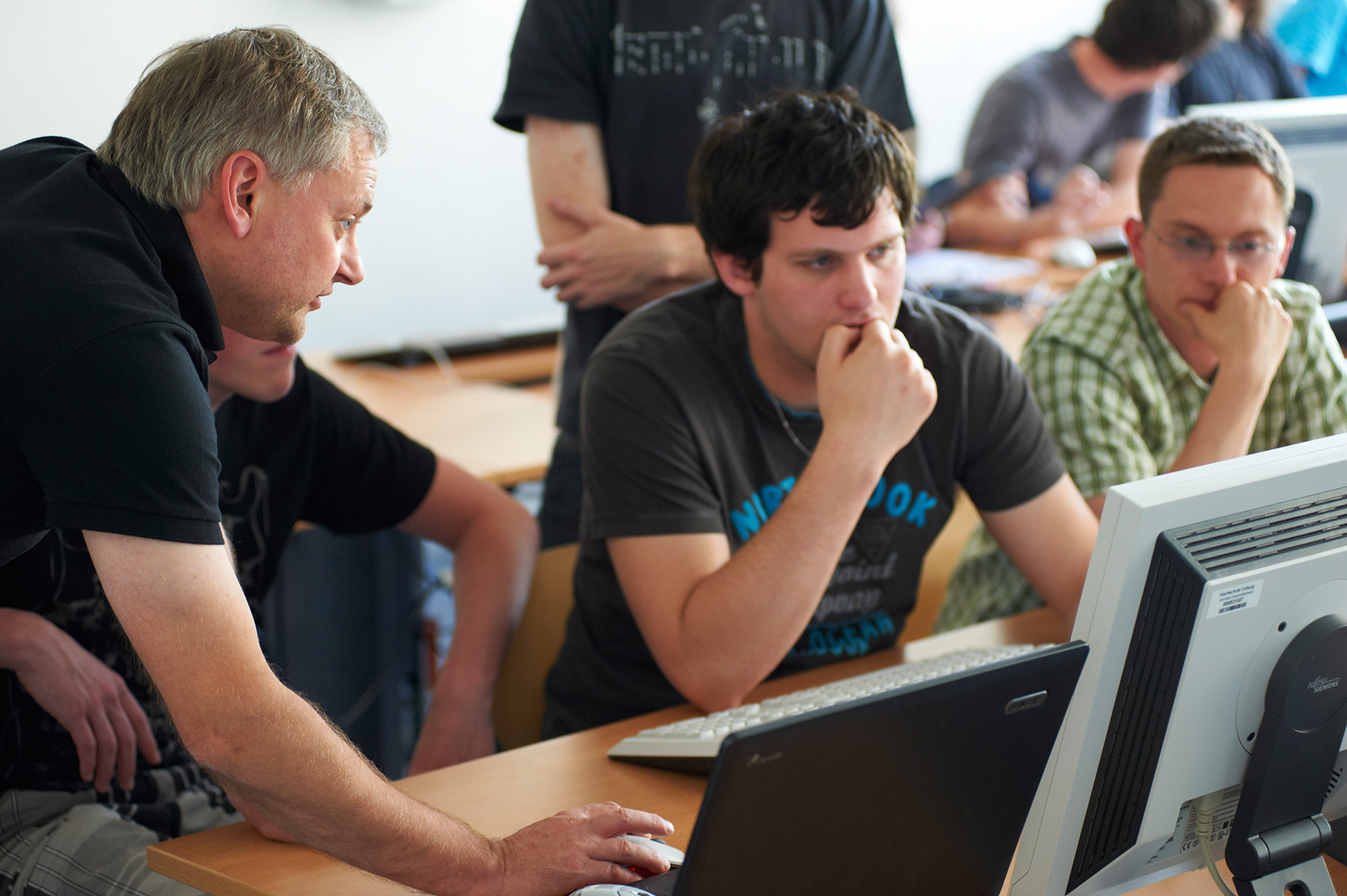 In einem Hörsaal der Hochschule Coburg konzentriert sich eine Gruppe auf einen Computerbildschirm. Ein älterer Mann deutet auf den Monitor, während drei jüngere Personen von ihren Schreibtischen aus aufmerksam zuschauen. In eine lebhafte Diskussion oder Unterrichtsstunde vertieft, verkörpern sie den Geist des kollaborativen Lernens.