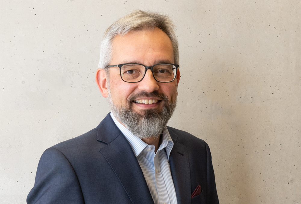 A man with gray hair and a beard, wearing glasses and a dark suit, smiles at the camera. He stands against a neutral, light-colored background, exuding an air of distinguished academia often associated with Hochschule Coburg.