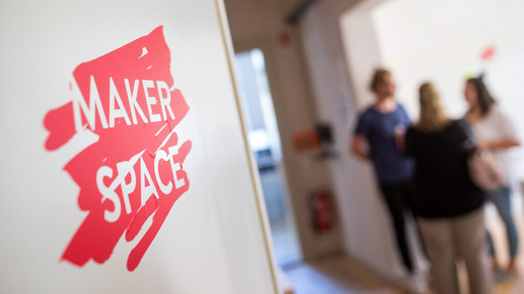 An einer Wand der Hochschule Coburg prangt ein Schild mit der Aufschrift „Maker Space“. Im unscharfen Hintergrund sind drei Personen zu sehen, die sich in einem hell erleuchteten Raum unterhalten.