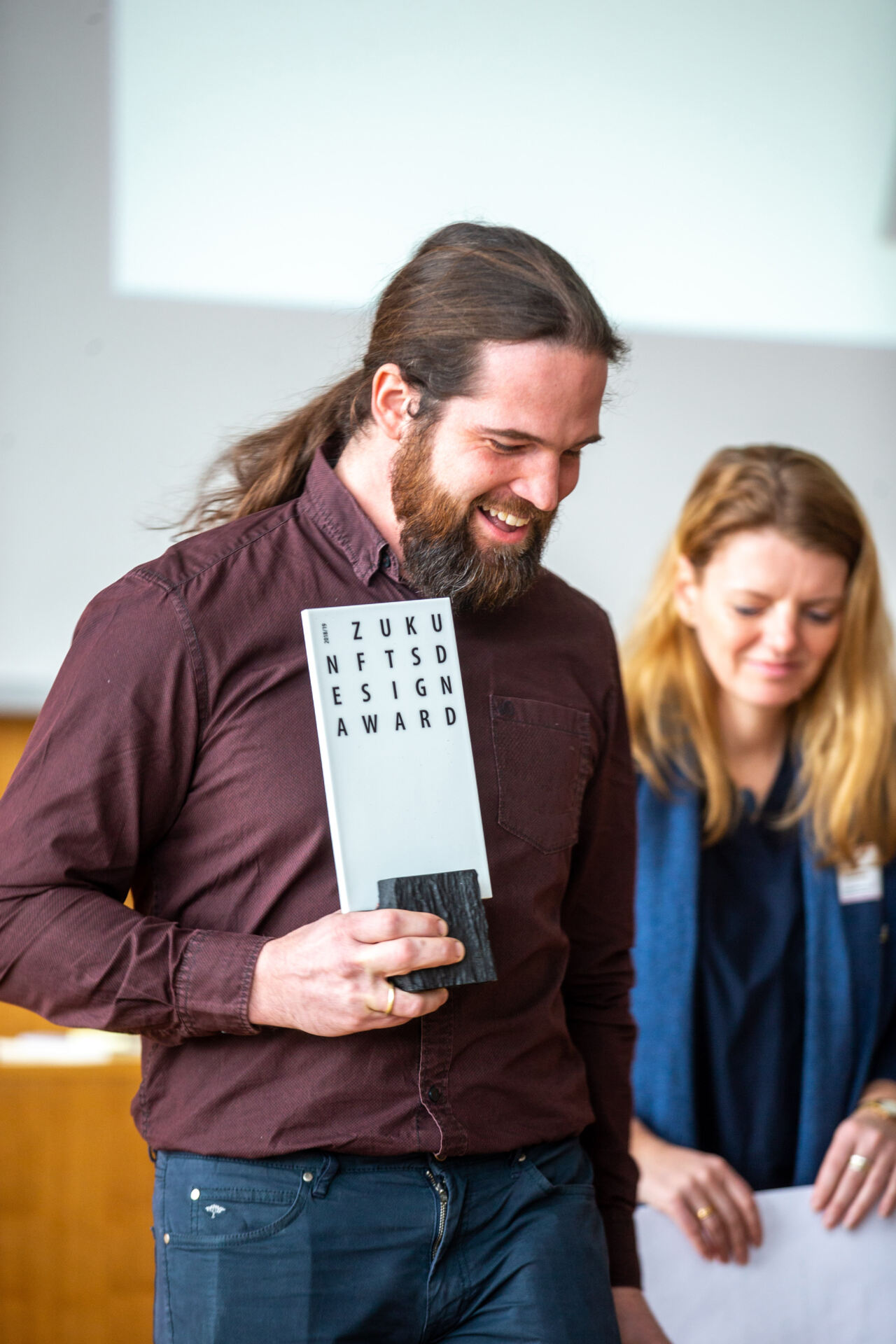 Ein bärtiger Mann lächelt und hält eine Plakette mit dem Designpreis der Hochschule Coburg in den Händen. Neben ihm steht eine lächelnde Frau in einem Raum mit unscharfem Hintergrund und Holzvertäfelung.