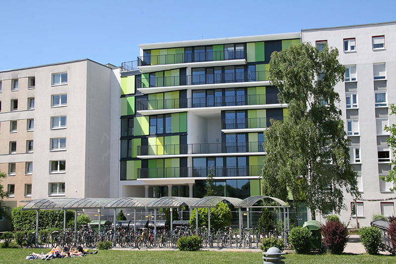 Ein modernes Wohngebäude mit grün-weißen Fassadenpaneelen in der Nähe der Hochschule Coburg ist von Bäumen umgeben. Im Vordergrund stehen Fahrräder unter einer Überdachung, während Menschen auf der Wiese unter einem klaren blauen Himmel ein Sonnenbad nehmen.