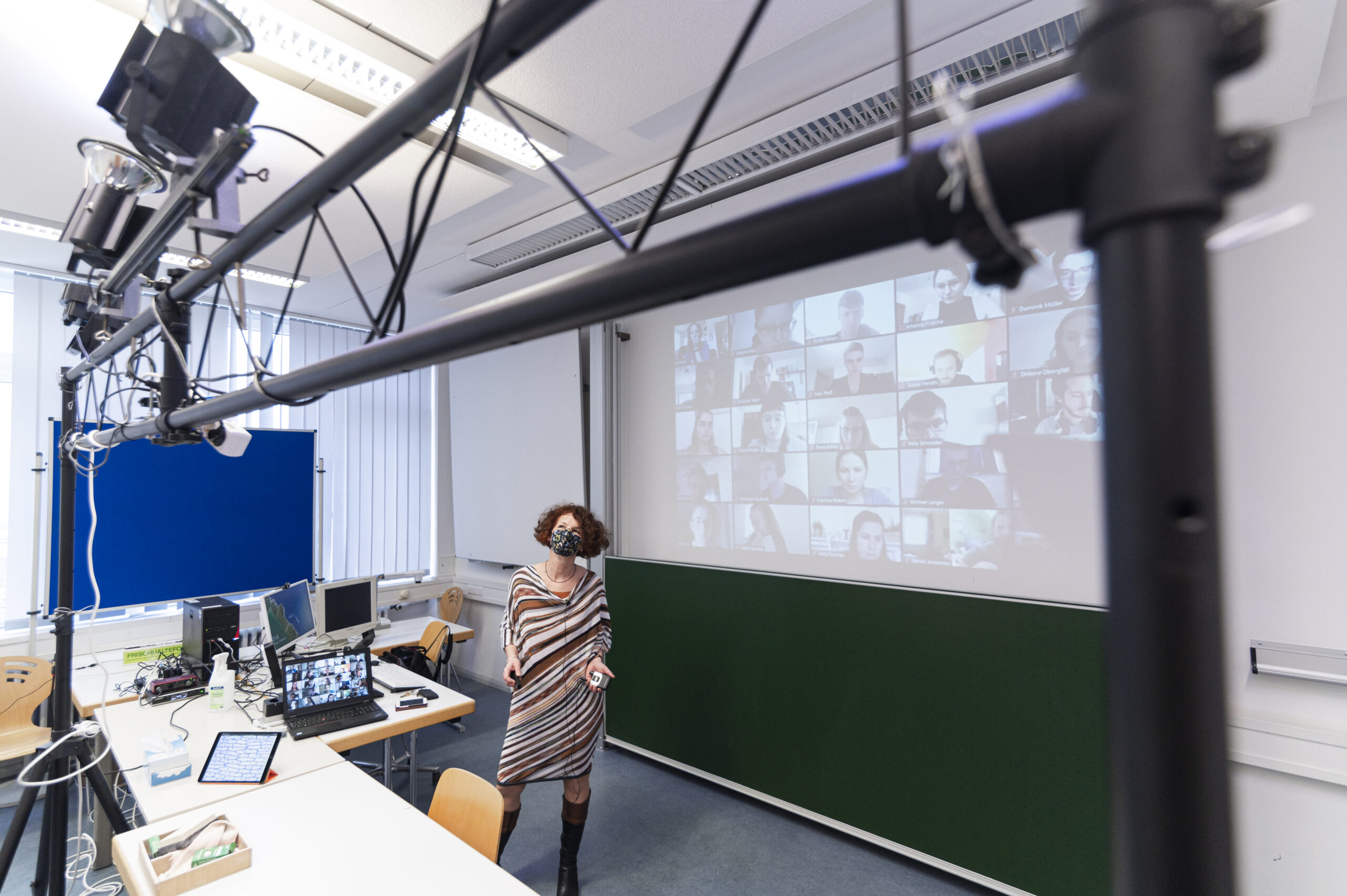 Eine Person steht in einem Hörsaal der Hochschule Coburg. Auf einem großen Bildschirm wird ein Raster der Teilnehmer einer Videokonferenz angezeigt. Der Raum ist mit Computern, Monitoren und technischer Ausrüstung ausgestattet. Im Vordergrund ist eine Kameraausrüstung zu sehen, die die dynamische Lernumgebung einfängt.