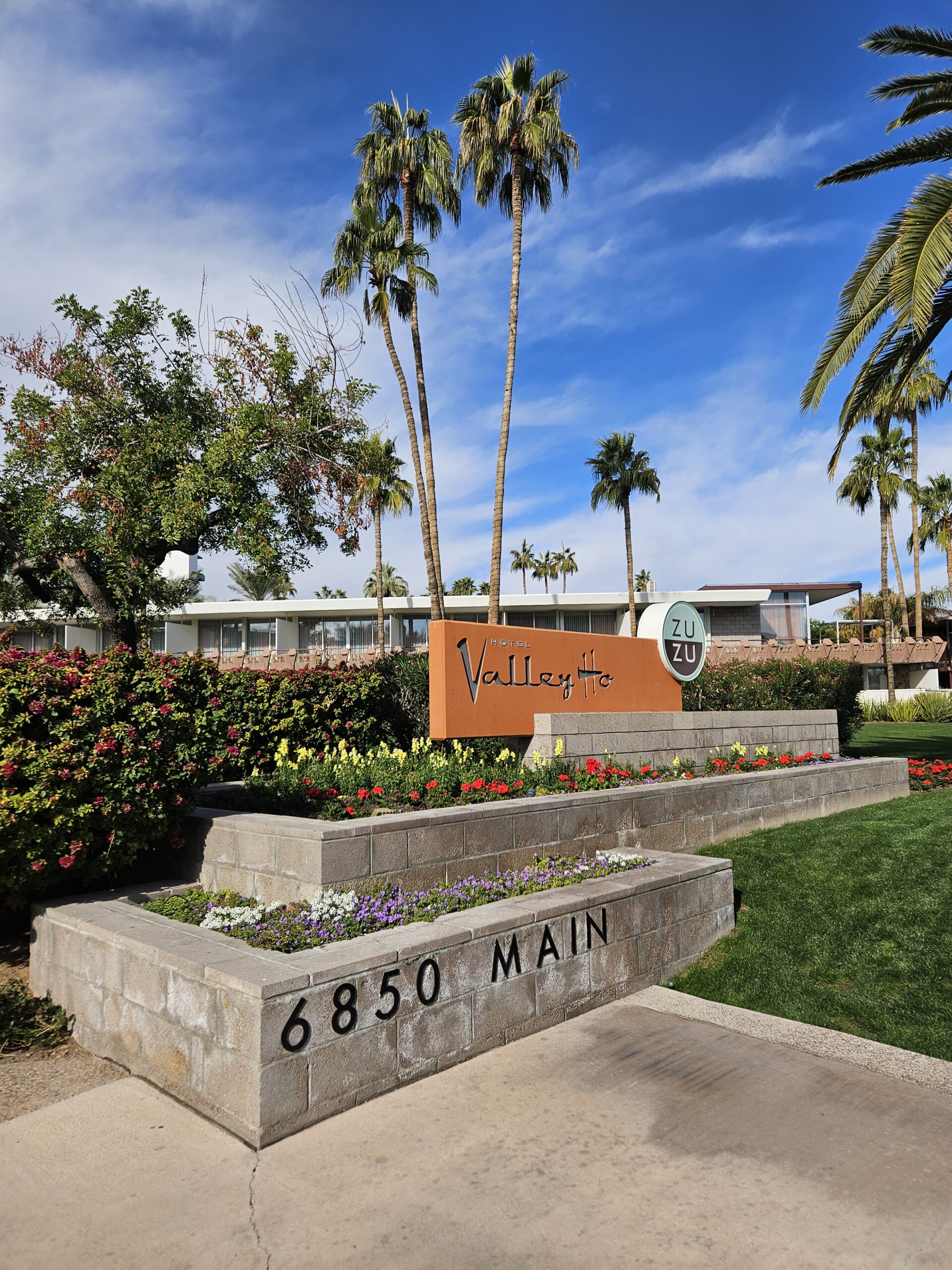 The image depicts a landscaped entrance to a building featuring a sign that reads "6850 Main" alongside the names "Valley Ho" and "ZuZu" on a circular sign. The scene is enhanced by palm trees, flowering plants, and the vibrant blue sky reminiscent of Hochschule Coburg's inviting campus ambiance.