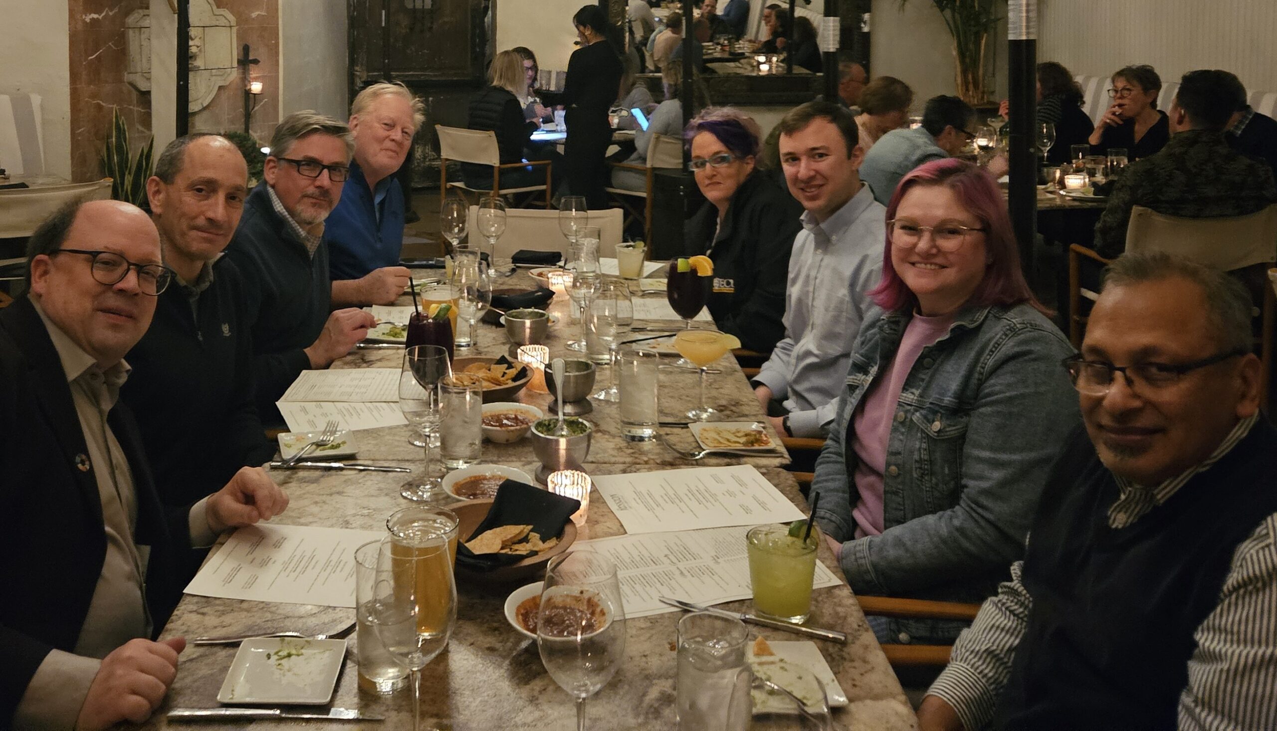 A group of eight people from Hochschule Coburg sit around a long dining table in a cozy, dimly lit restaurant. Menus, drinks, and appetizers adorn the table, creating a casual and inviting atmosphere for lively conversation.