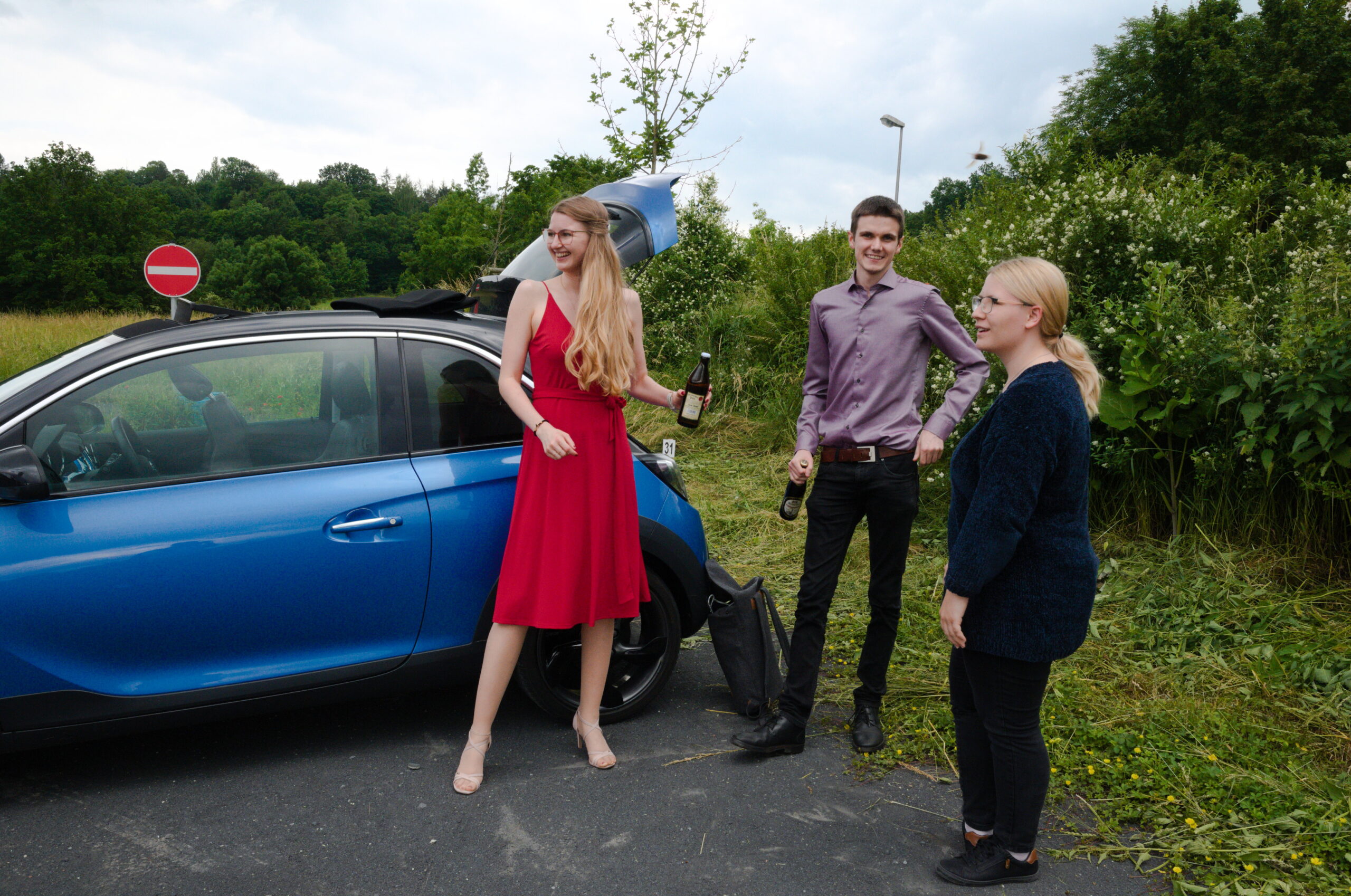 Drei Personen stehen neben einem blauen Auto auf einer Landstraße in der Nähe der Hochschule Coburg. Eine Frau in einem roten Kleid und High Heels hält lächelnd eine Flasche. Zwei weitere, ein Mann in einem lila Hemd und eine Frau in Schwarz, halten ebenfalls Flaschen, umgeben von üppigem Grün.