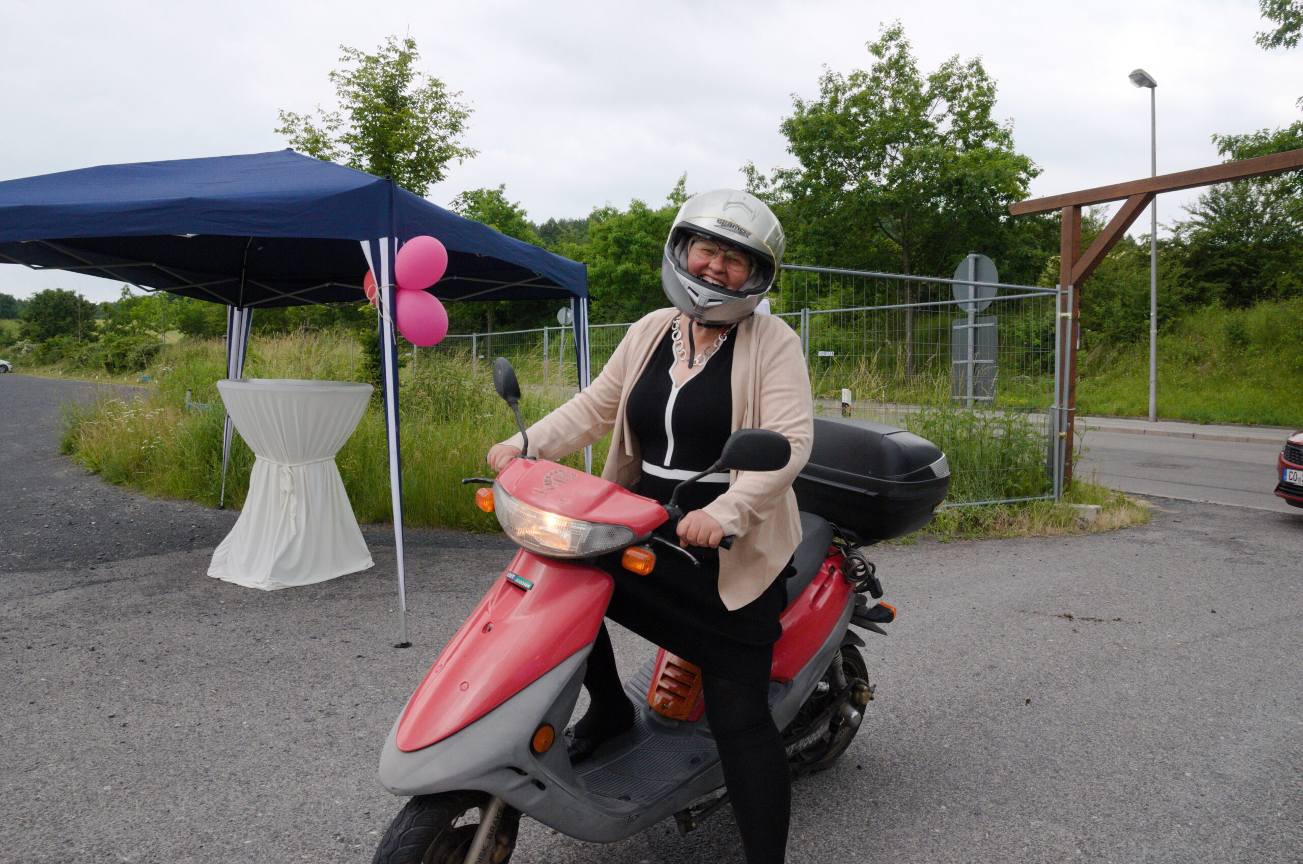 Eine Person mit Helm und schwarzer Jacke fährt mit einem roten Roller auf einer asphaltierten Straße in der Nähe der Hochschule Coburg. Im Hintergrund ist ein Pop-up-Zelt mit rosa Luftballons und grünem Gras zu sehen.