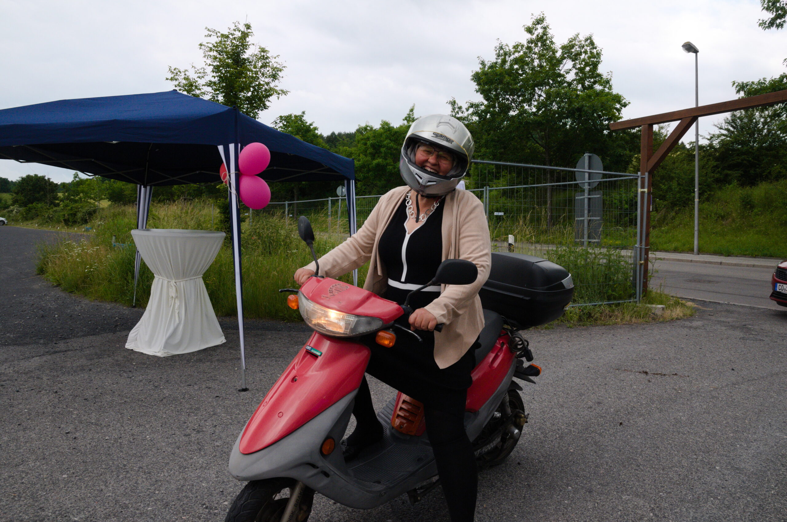 Eine Person mit Helm und beiger Jacke fährt auf einem roten Roller durch das Außengelände der Hochschule Coburg. Im Hintergrund ziert ein mit rosa Luftballons geschmückter Baldachin und ein runder Stehtisch das üppige Grün, das das Gelände umgibt.