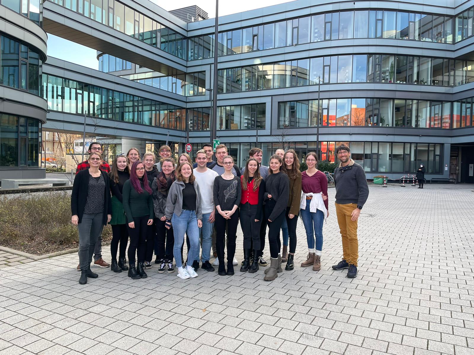 Auf einem gepflasterten Hof vor einem modernen Glasbau der Hochschule Coburg steht eine Gruppe von Menschen zusammen. Sie sind leger gekleidet, der Himmel ist klar.