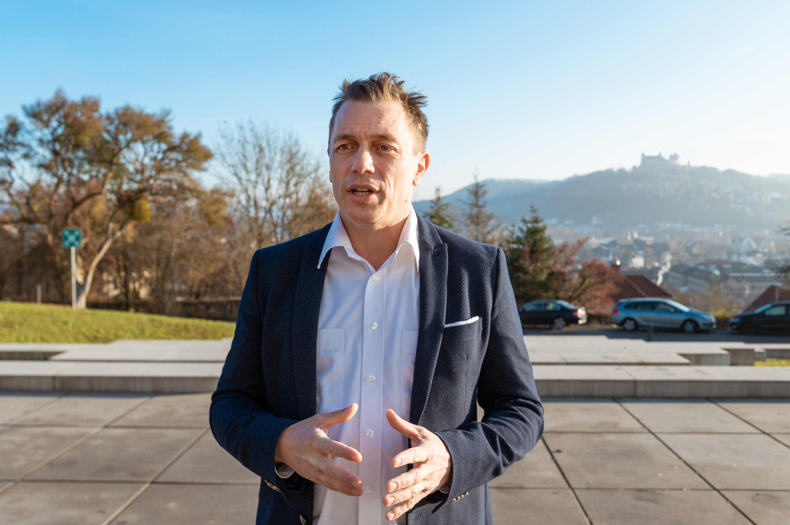 Ein Mann in weißem Hemd und dunkler Jacke steht draußen und gestikuliert mit den Händen. Hinter ihm ist der malerische Blick auf eine Stadtlandschaft mit Hügeln und einer Burg unter dem klaren blauen Himmel zu sehen, der an Aussichten in der Nähe der Hochschule Coburg erinnert.