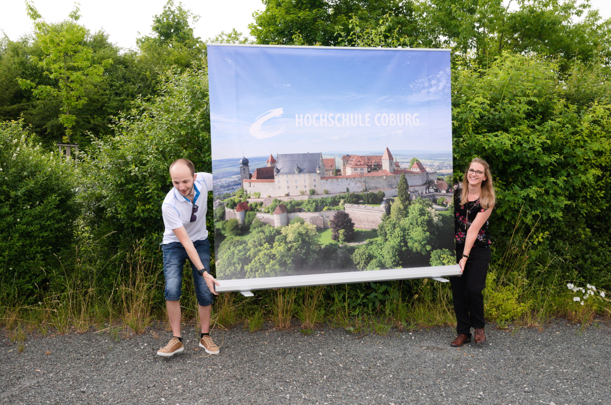 Zwei Personen halten stolz ein großes Banner mit einer malerischen Aussicht auf ein historisches Schloss und der Aufschrift „Hochschule Coburg“. Sie lächeln inmitten üppigen Grüns und fangen die Essenz dieses akademischen Schatzes in einer bezaubernden Außenumgebung ein.